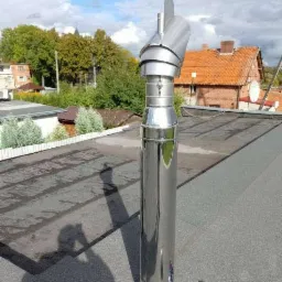 Błyszczący, stalowy wkład kominowy z ozdobną wiatrowskazową nakładką na dachu pokrytym papą, obok leżące narzędzia.