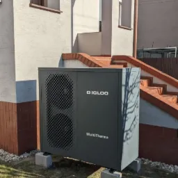 Szara pompa ciepła marki Igloo MultiTherma, zamontowana na betonowych bloczkach przed budynkiem mieszkalnym z ceglanymi elementami.