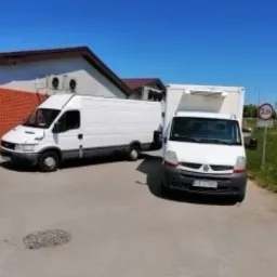 Dwa białe busy dostawcze zaparkowane obok siebie na parkingu, jeden z zabudową kontenerową, drugi typu furgon, w tle budynek z klimatyzacją i znak drogowy.