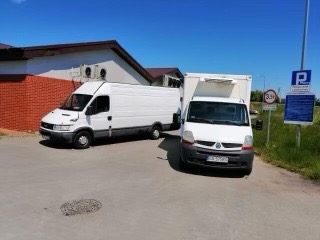 Dwa białe busy dostawcze zaparkowane obok siebie na parkingu, jeden z zabudową kontenerową, drugi typu furgon, w tle budynek z klimatyzacją i znak drogowy.