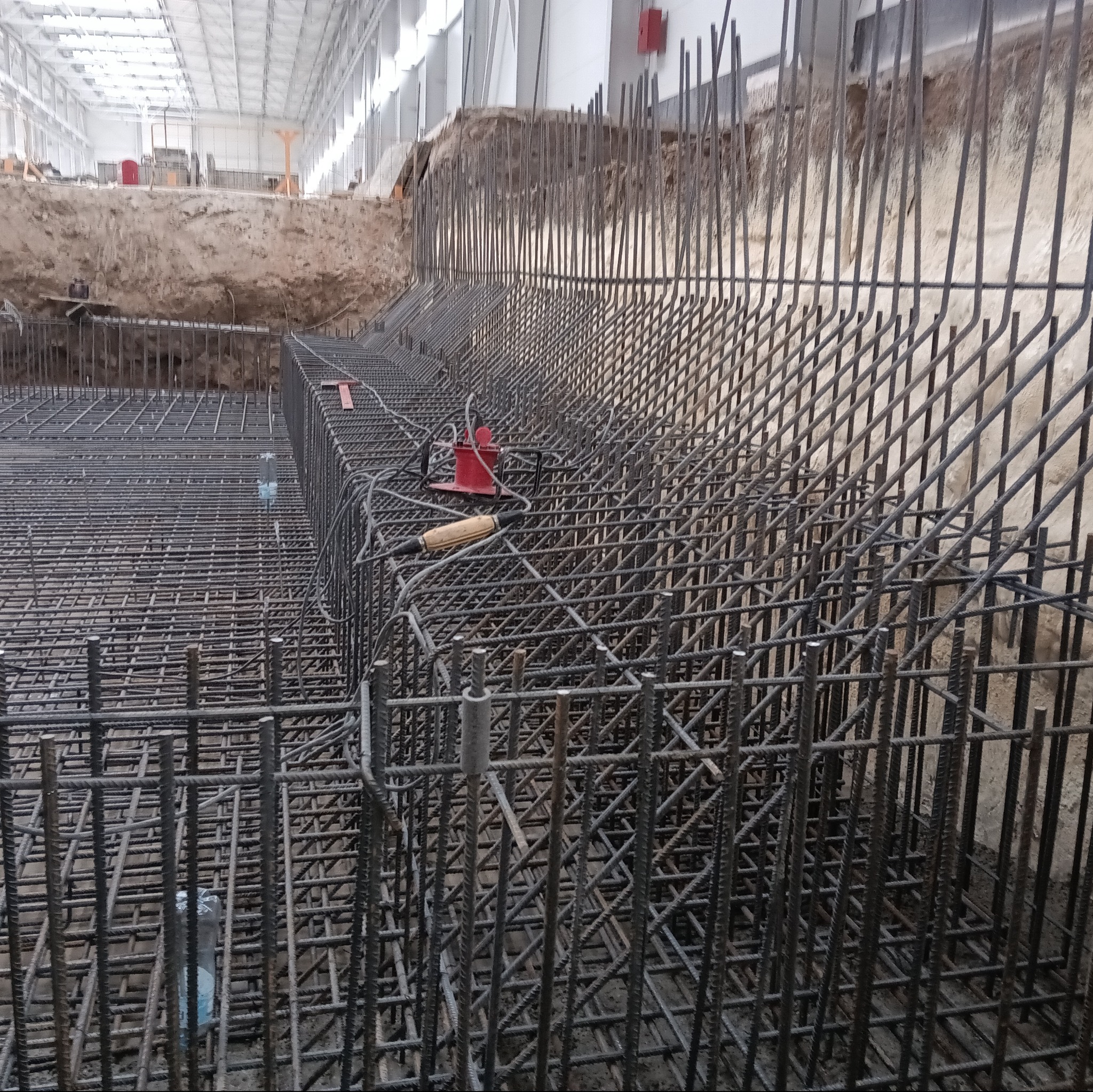 Siatka zbrojeniowa fundamentów w wykopie, widoczny sprzęt do łączenia prętów, w tle ściana wykopu i hala.