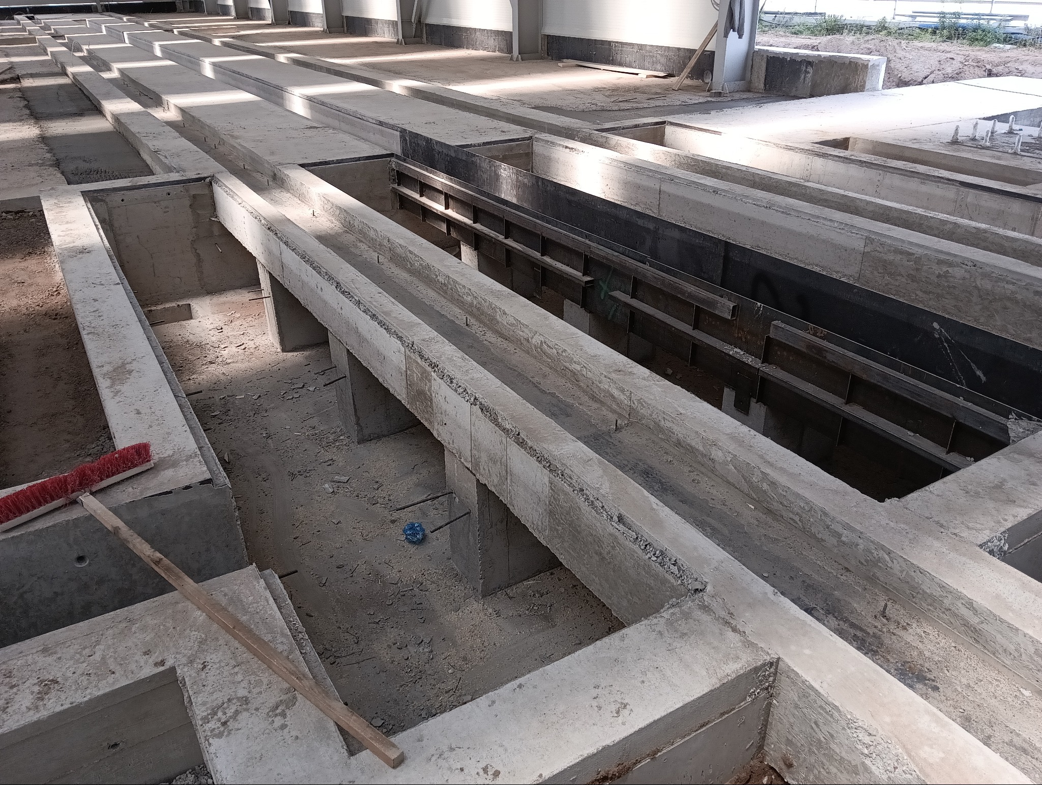 Surowy stan fundamentów z widocznymi betonowymi belkami i metalowymi elementami konstrukcyjnymi, uzupełniony czerwoną szczotką do zamiatania na pierwszym planie.