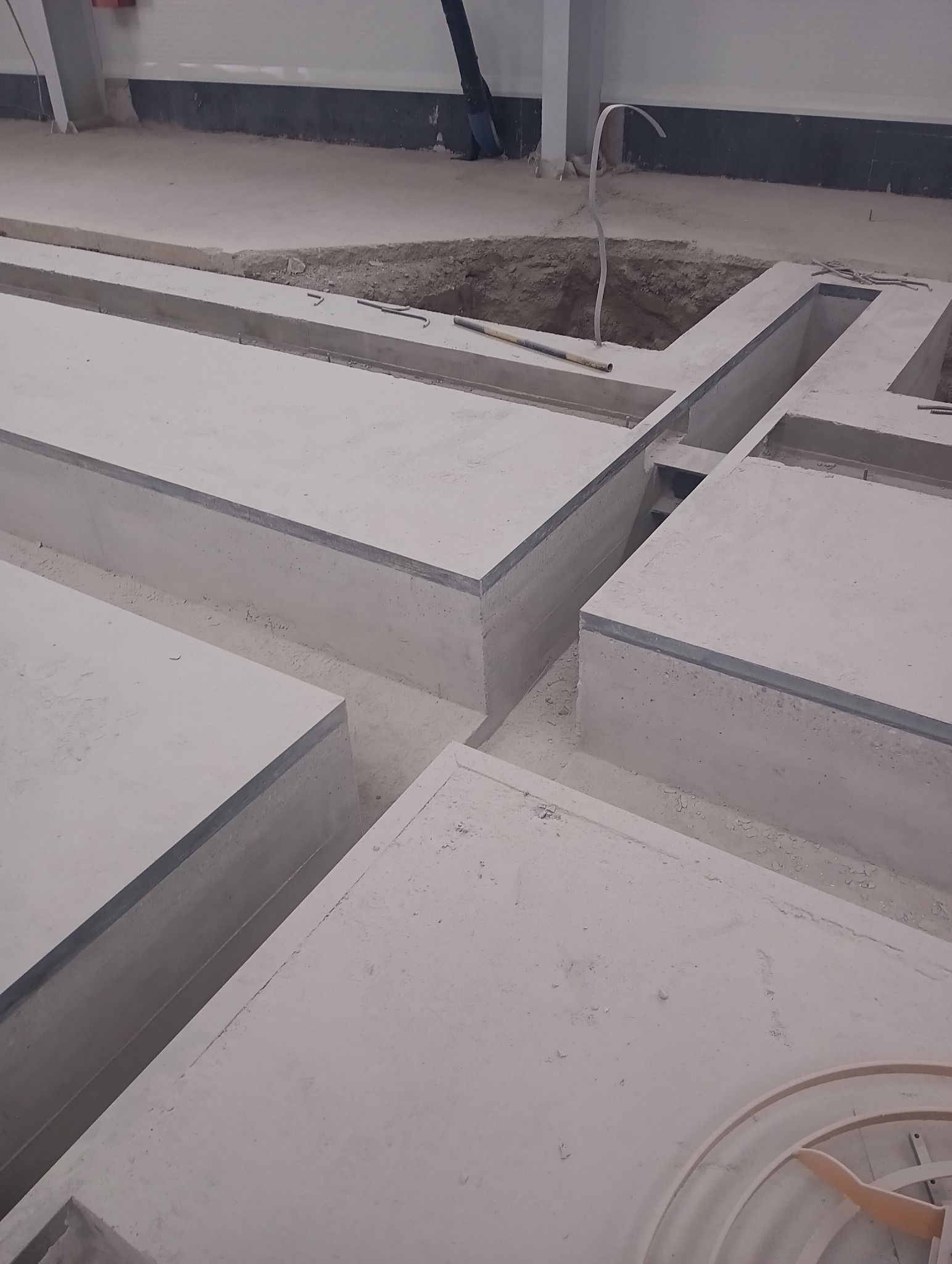 Surowe, betonowe fundamenty z wykopem w tle, widoczne narzędzia budowlane i kable. Wnętrze hali.