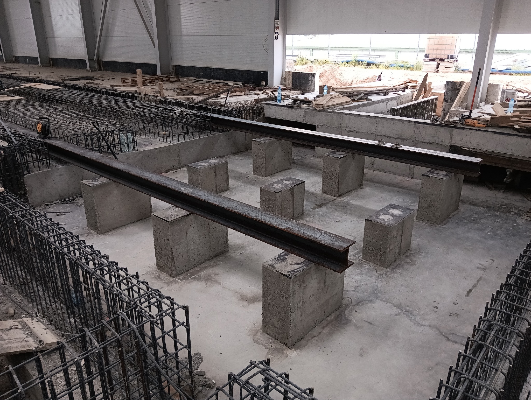 Przygotowanie fundamentów pod konstrukcję, widoczne betonowe bloczki, stalowe belki i zbrojenie w dużej hali.