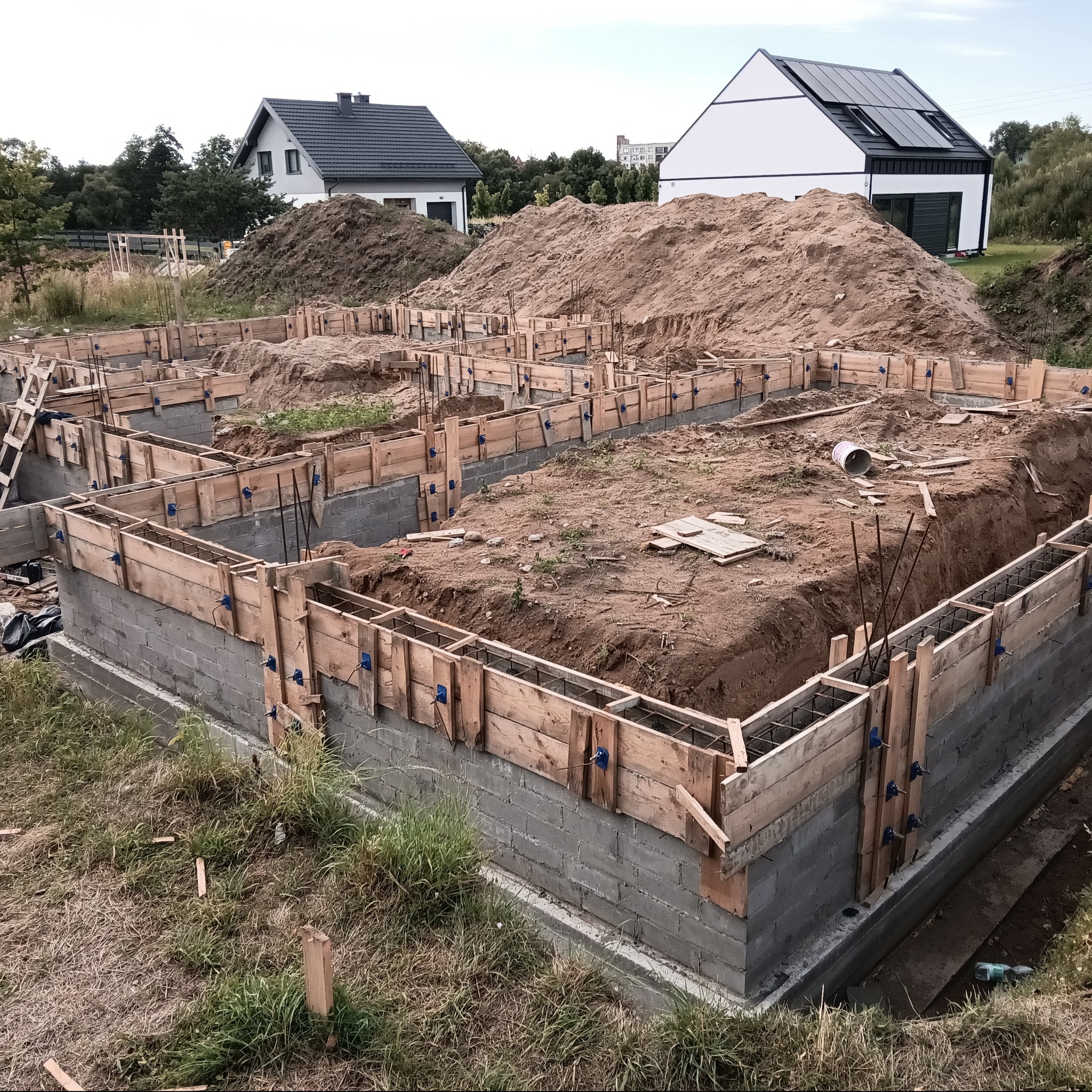Wykopane fundamenty pod budowę domu, zbrojenie w szalunkach drewnianych, w tle dwa domy jednorodzinne i hałda ziemi.