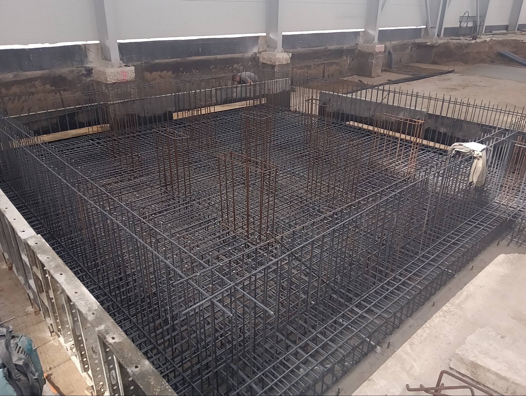 Siatka zbrojeniowa fundamentu w wykopie, widoczny pracownik w tle, przygotowana do zalania betonem.