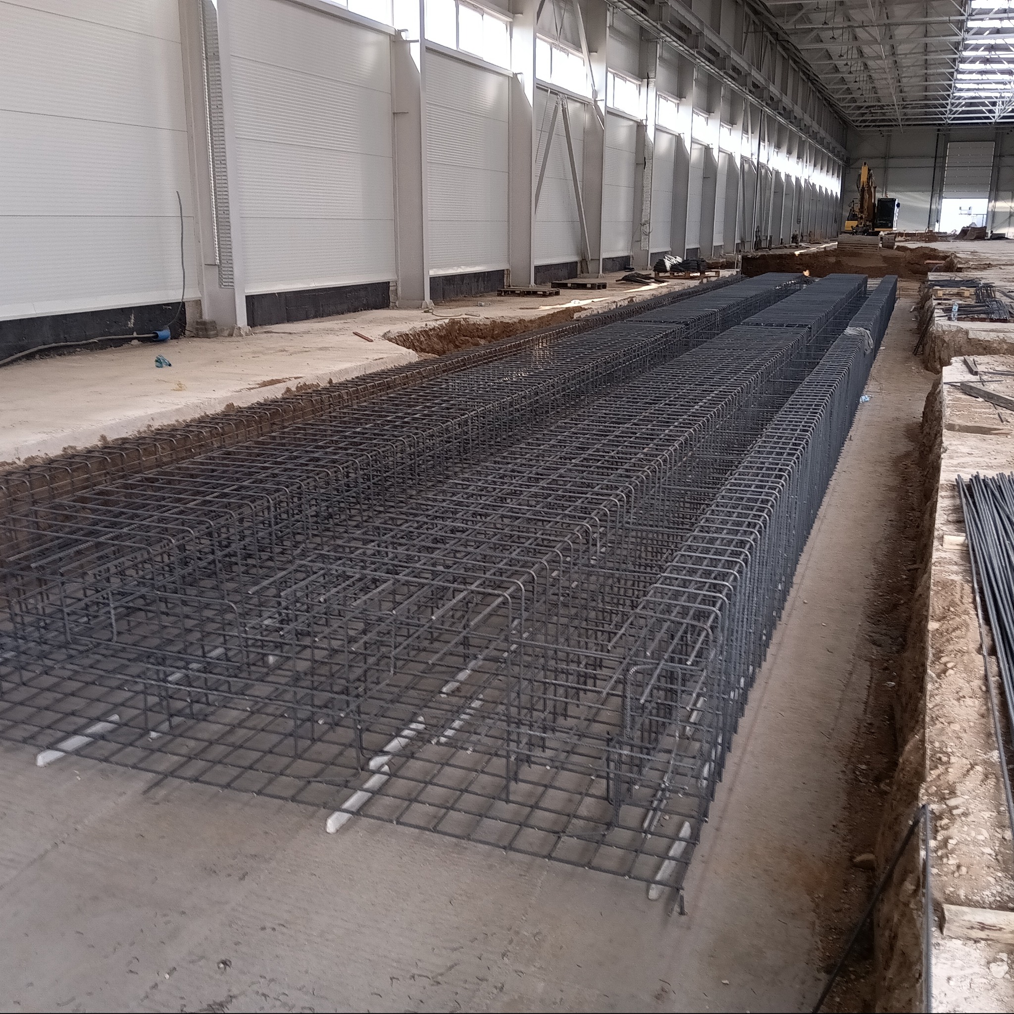 Siatka zbrojeniowa fundamentów ułożona w wykopie na hali, widoczne białe elementy dystansowe pod zbrojeniem oraz równe ułożenie prętów.