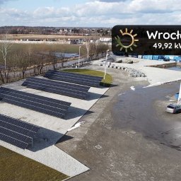 Panele - Instalacja paneli słonecznych o mocy 49,92 kWp we Wrocławiu, widok z góry na rząd paneli na szarym żwirze, z budynkiem przemysłowym w tle i zaparkowanymi samochodami.