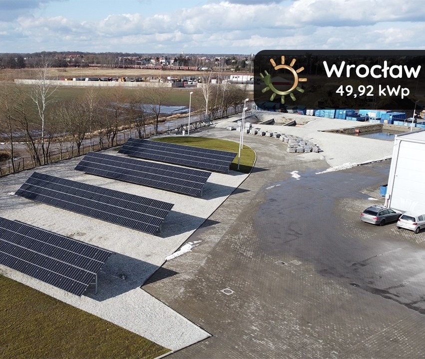 Instalacja paneli słonecznych o mocy 49,92 kWp we Wrocławiu, widok z góry na rząd paneli na szarym żwirze, z budynkiem przemysłowym w tle i zaparkowanymi samochodami.