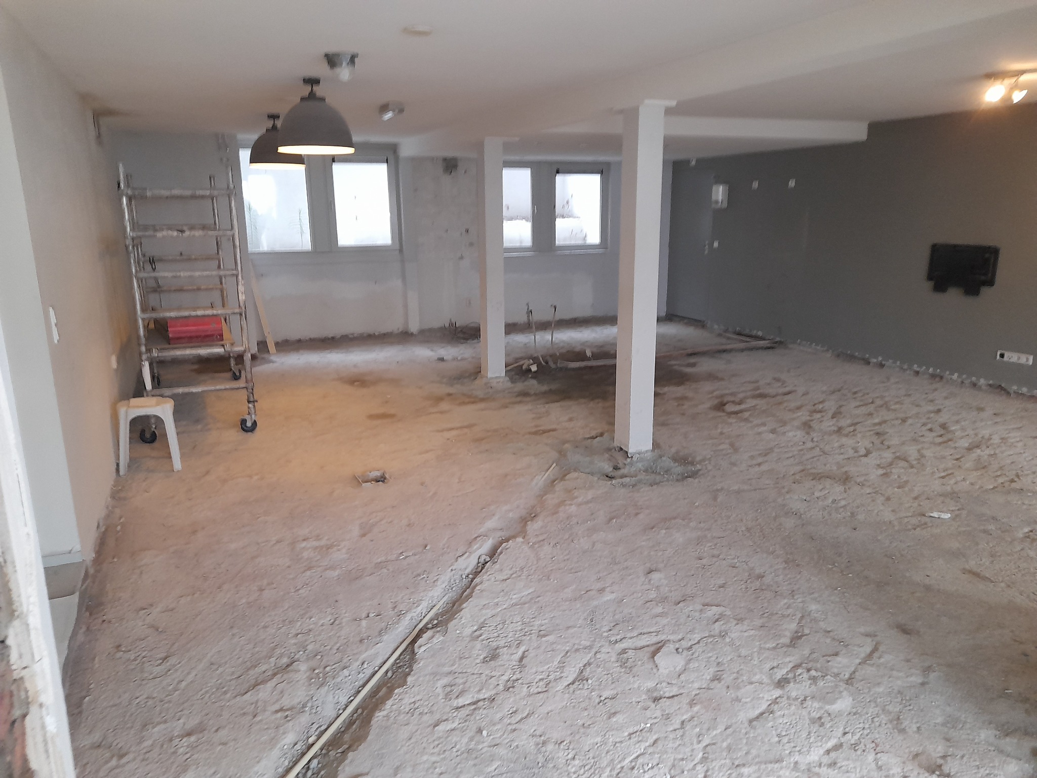 Wnętrze w trakcie prac remontowych: zerwana podłoga, odsłonięte instalacje, drabina aluminiowa z czerwoną skrzynką narzędziową, widoczne ślady po skuwaniu płytek i tapet, przygotowanie do dalszych...