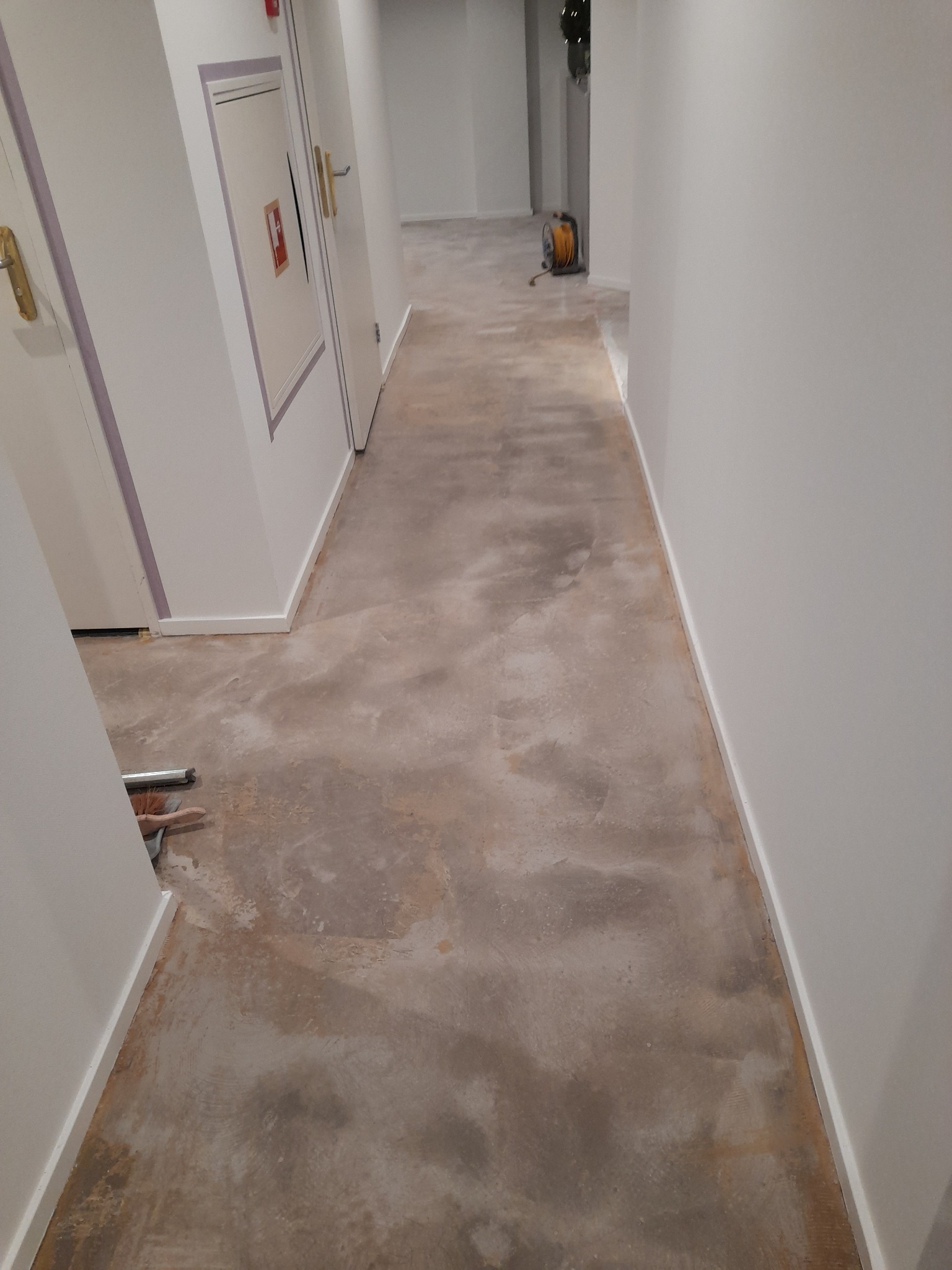 Korytarz w trakcie prac przygotowawczych do ułożenia podłogi, widoczne resztki starej wylewki i narzędzia przy ścianie.