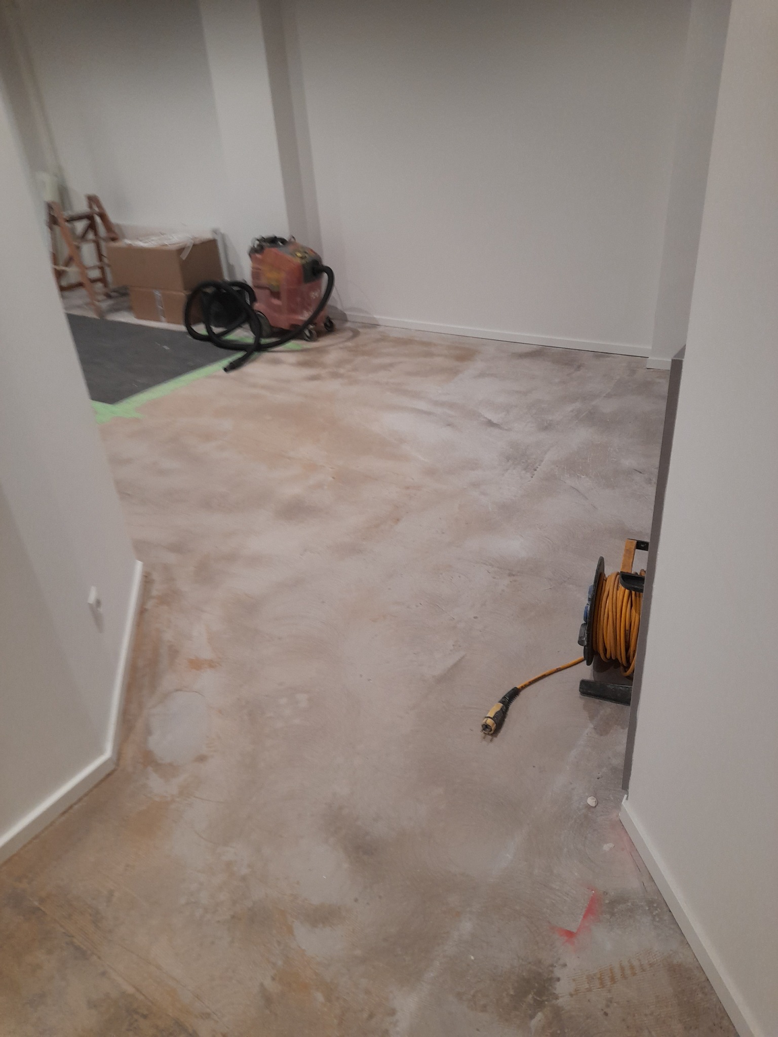 Przygotowanie podłoża w pomieszczeniu: wyrównana, szlifowana posadzka z widocznymi śladami po wcześniejszych pracach, w tle drabina, odkurzacz przemysłowy i kartony, na pierwszym planie zwinięty...