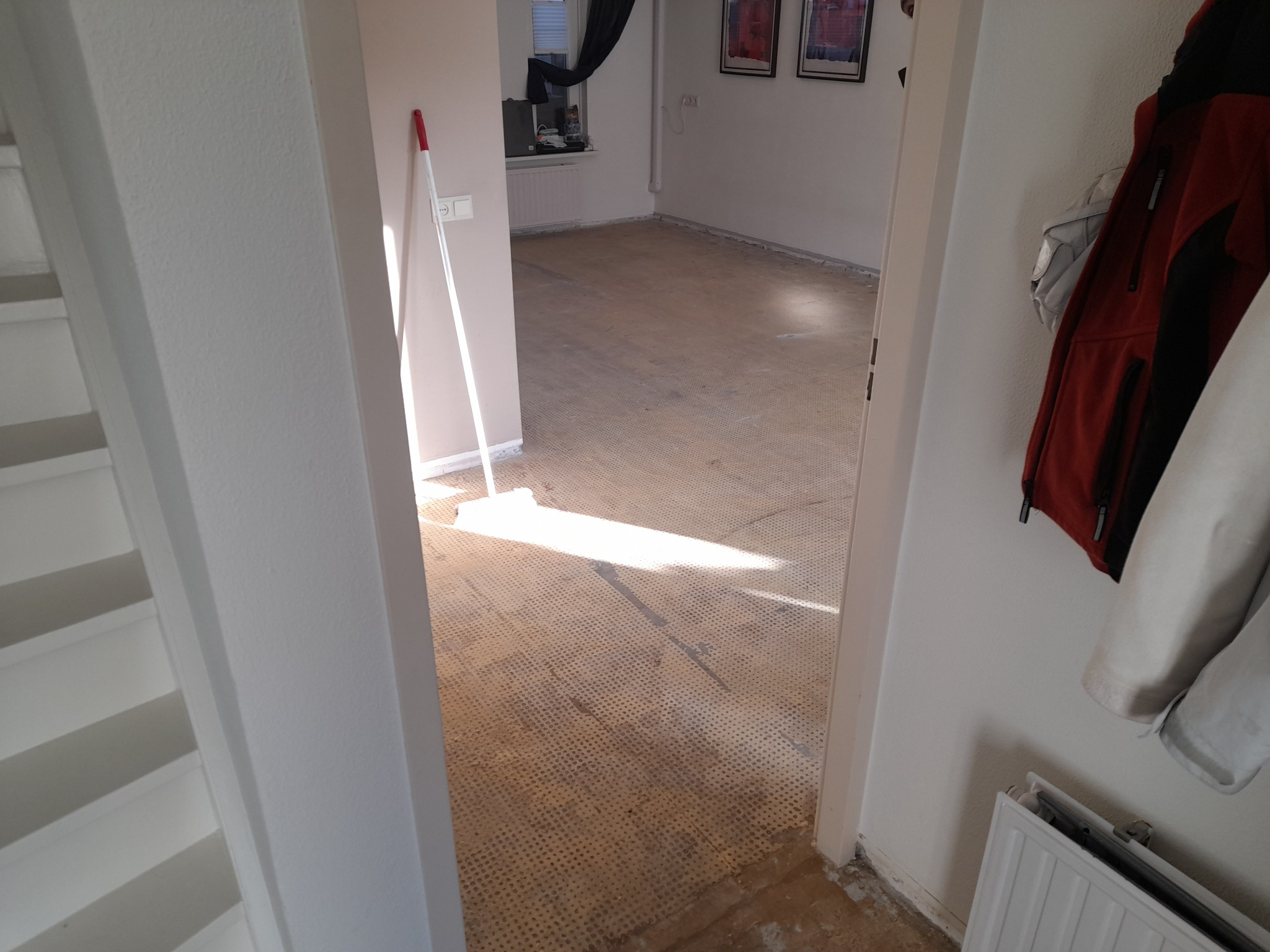 Widok przez drzwi na pomieszczenie po usunięciu podłogi, widoczne ślady po kleju i dziurki, w rogu oparta miotła, na ścianie wiszą ubrania.