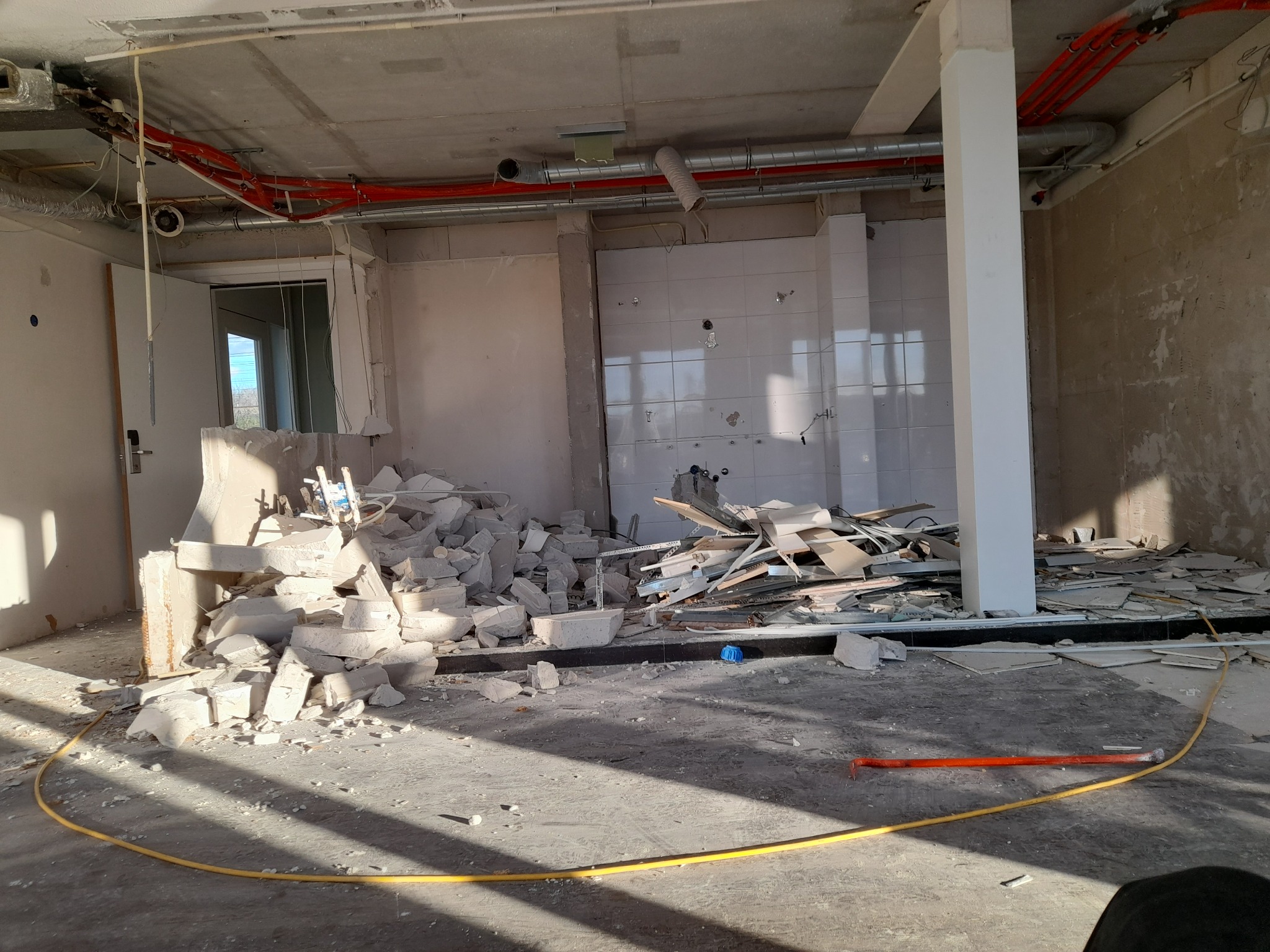 Wnętrze po demontażu: gruz, fragmenty płytek i instalacji, widoczne rury pod sufitem, resztki ścian i wyposażenia w hotelowym pokoju.