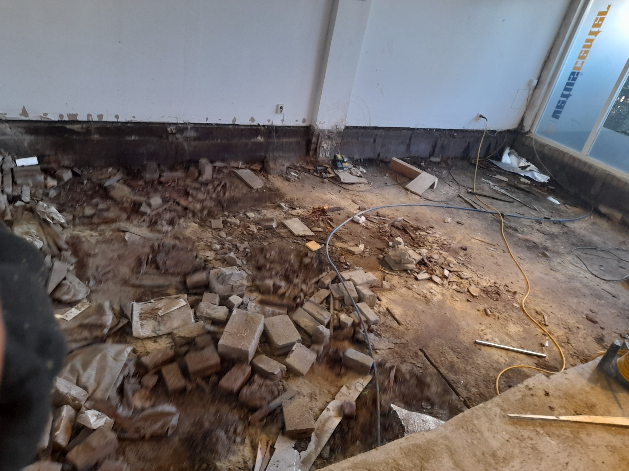 Wnętrze budynku w trakcie rozbiórki: gruz, fragmenty cegieł i przewodów na podłodze, ślady po usuniętych elementach przy ścianie.