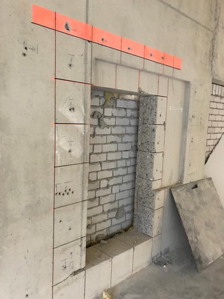 Wykucie otworu w betonowej ścianie z widoczną cegłą, zaznaczone linie cięcia i pomarańczowa listwa kontrolna.