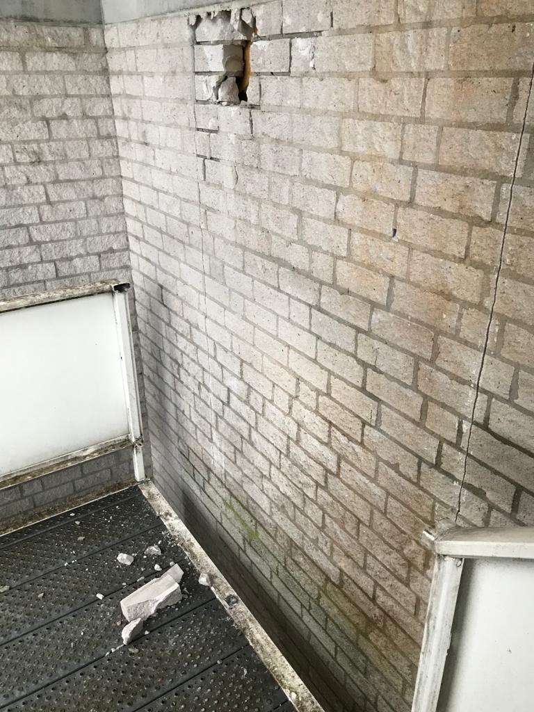 Uszkodzona ściana z cegły z widocznym ubytkiem i pęknięciami, gruz leżący na podłodze, perspektywa z dołu.