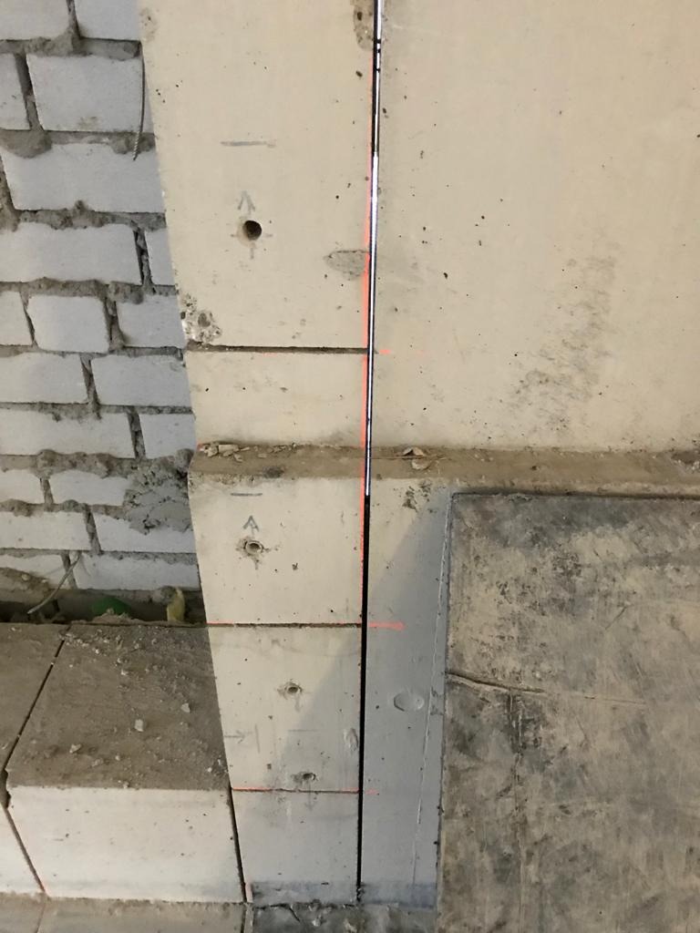 Wewnętrzna ściana budynku w trakcie rozbiórki, widoczne bloki betonowe z otworami i czerwoną linią lasera, fragment ściany z cegły oraz betonowa podłoga.