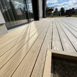 Nowoczesny taras z desek kompozytowych w odcieniach beżu, z widocznym fragmentem obudowy rabaty i dużym oknem domu odbijającym błękitne niebo z chmurami.