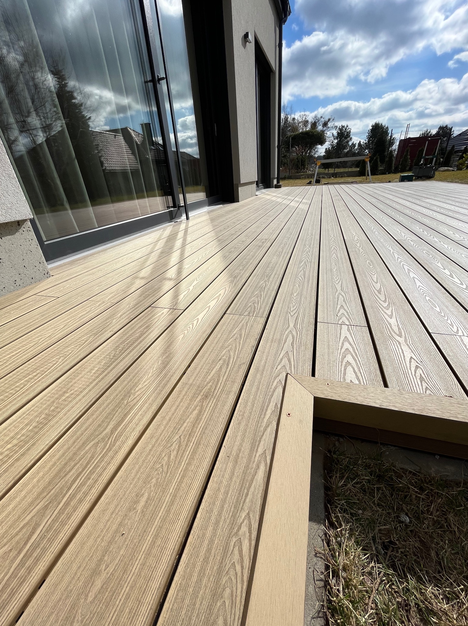 Nowoczesny taras z desek kompozytowych w odcieniach beżu, z widocznym fragmentem obudowy rabaty i dużym oknem domu odbijającym błękitne niebo z chmurami.