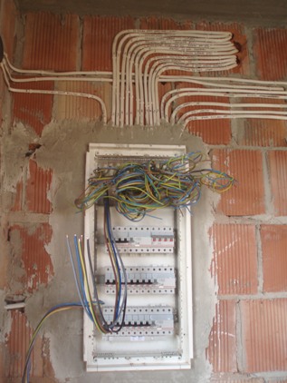 Rozdzielnica elektryczna w stanie surowym, widoczne przewody wychodzące z listwy i prowadzone w peszlach po murze z czerwonej cegły.