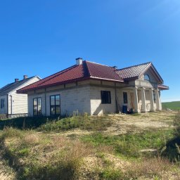 J.B. SPÓŁKA Z OGRANICZONĄ ODPOWIEDZIALNOŚCIĄ - Dom w trakcie budowy z czerwoną dachówką i kolumnami przy wejściu, na tle błękitnego nieba i zielonego pola. Widoczny stan surowy otwarty, z elementami wykończeniowymi dachu.