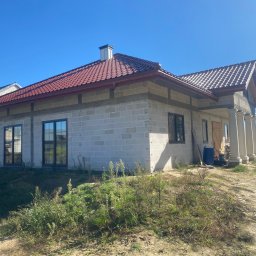 J.B. SPÓŁKA Z OGRANICZONĄ ODPOWIEDZIALNOŚCIĄ - Niedokończony dom z czerwonym dachem i kolumnami, widok z zewnątrz w słoneczny dzień. Widoczne bloczki betonowe i elementy konstrukcyjne.