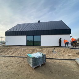 Nowoczesny dom z czarnym dachem z blachy na etapie wykończenia terenu. Widoczni pracownicy układający kostkę brukową. Paleta kostki na pierwszym planie.