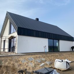 J.B. SPÓŁKA Z OGRANICZONĄ ODPOWIEDZIALNOŚCIĄ - Nowoczesny dom jednorodzinny z czarnym dachem i białymi ścianami w trakcie wykańczania terenu wokół budynku. Widoczne narzędzia i pracownicy w pomarańczowych ubraniach.