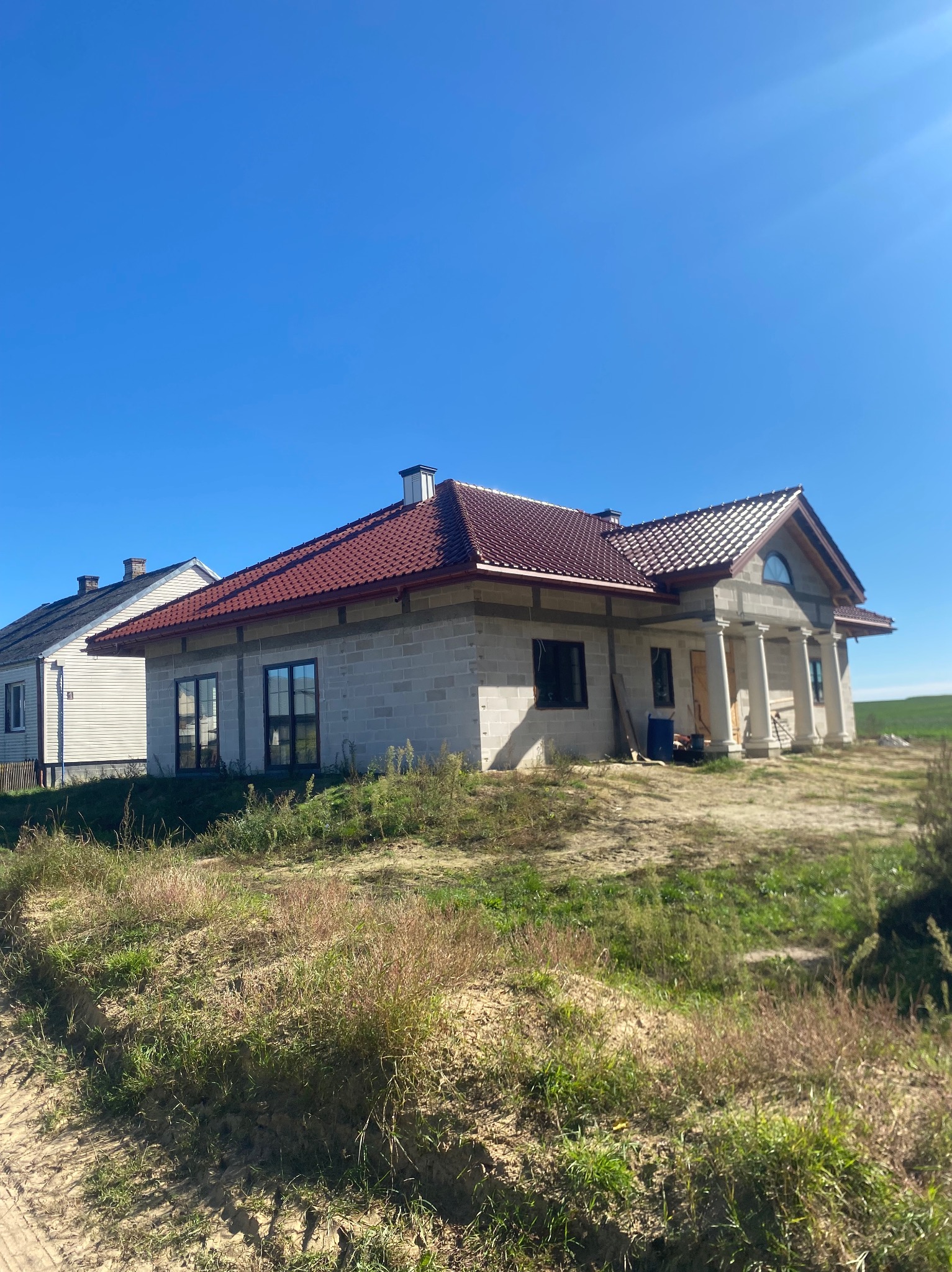 Dom w trakcie budowy z czerwoną dachówką i kolumnami przy wejściu, na tle błękitnego nieba i zielonego pola. Widoczny stan surowy otwarty, z elementami wykończeniowymi dachu.