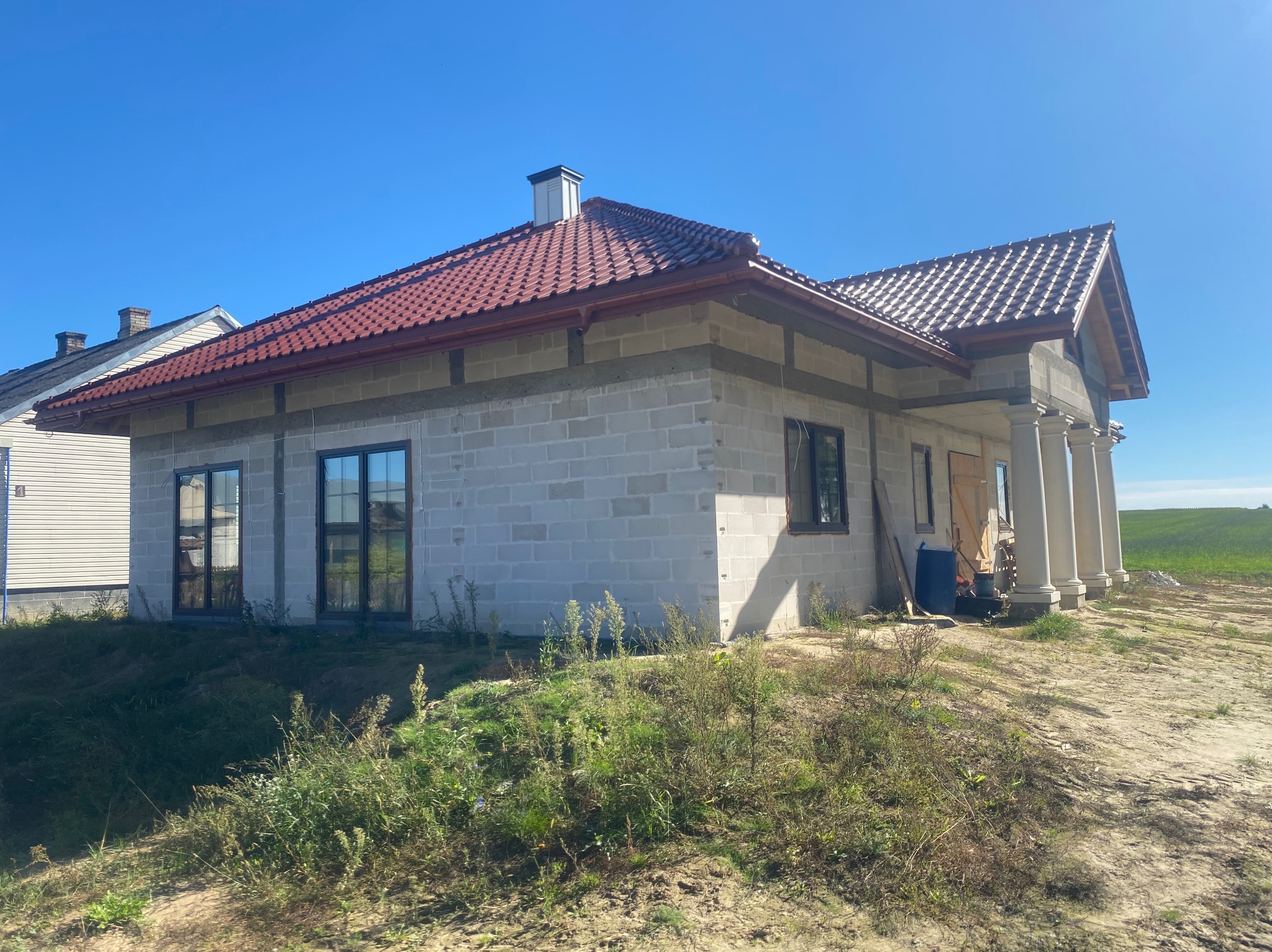 Niedokończony dom z czerwonym dachem i kolumnami, widok z zewnątrz w słoneczny dzień. Widoczne bloczki betonowe i elementy konstrukcyjne.