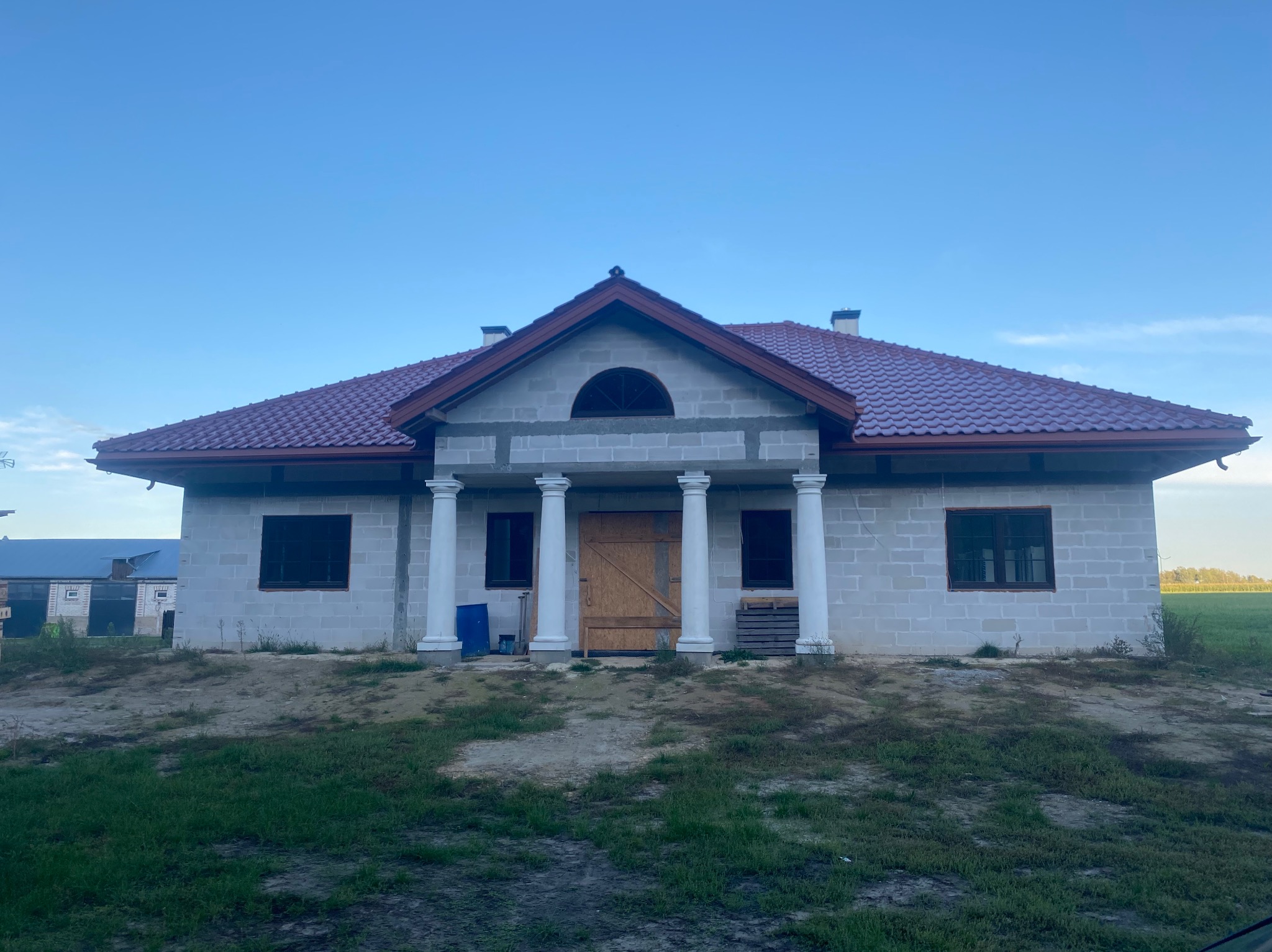 Dom w trakcie budowy z czerwonym dachem, białymi kolumnami i drewnianymi drzwiami. Widoczne bloczki betonowe i elementy konstrukcyjne. Krajobraz wiejski w tle.