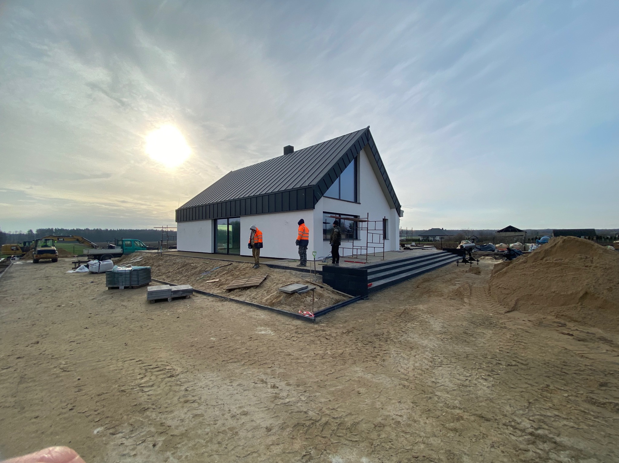 Nowoczesny dom jednorodzinny z ciemnym dachem dwuspadowym i dużymi oknami. Na pierwszym planie pracownicy w kamizelkach odblaskowych. Teren wokół domu w trakcie zagospodarowania.