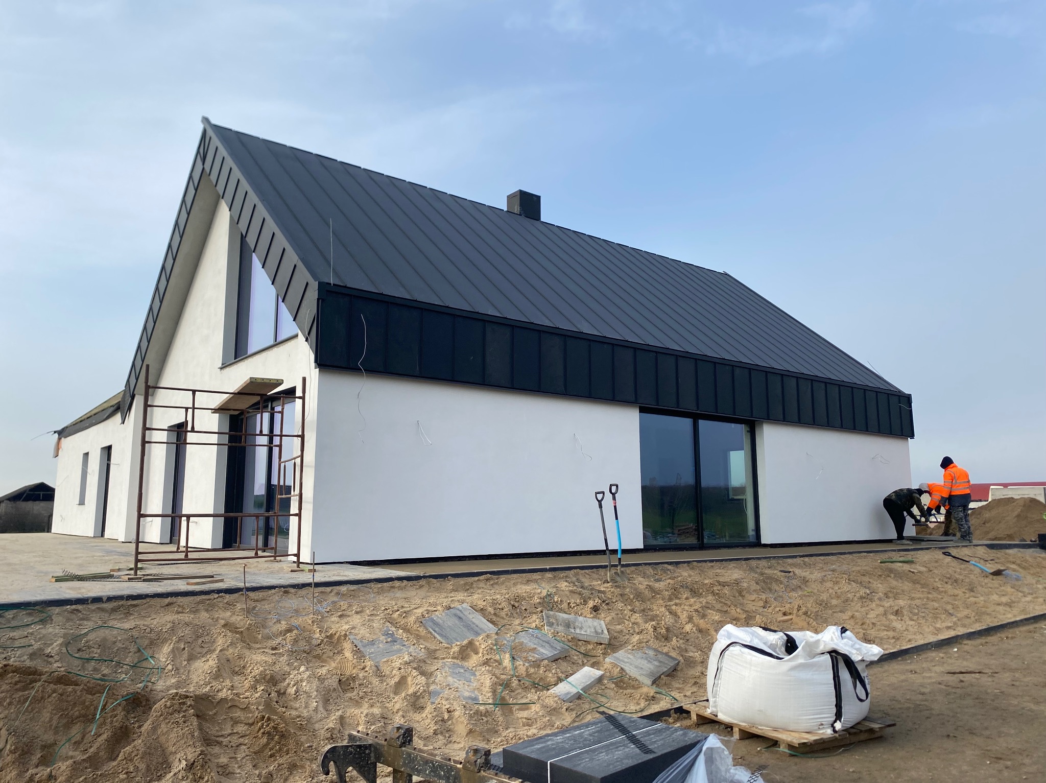 Nowoczesny dom jednorodzinny z czarnym dachem i białymi ścianami w trakcie wykańczania terenu wokół budynku. Widoczne narzędzia i pracownicy w pomarańczowych ubraniach.