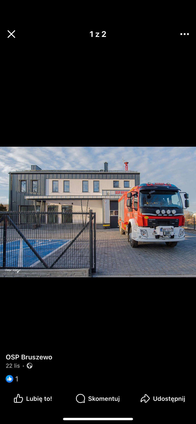 Budynek remizy strażackiej z szarym dachem i czerwonym wozem strażackim na zewnątrz. Widoczny parking z niebieskim oznaczeniem dla osób niepełnosprawnych.