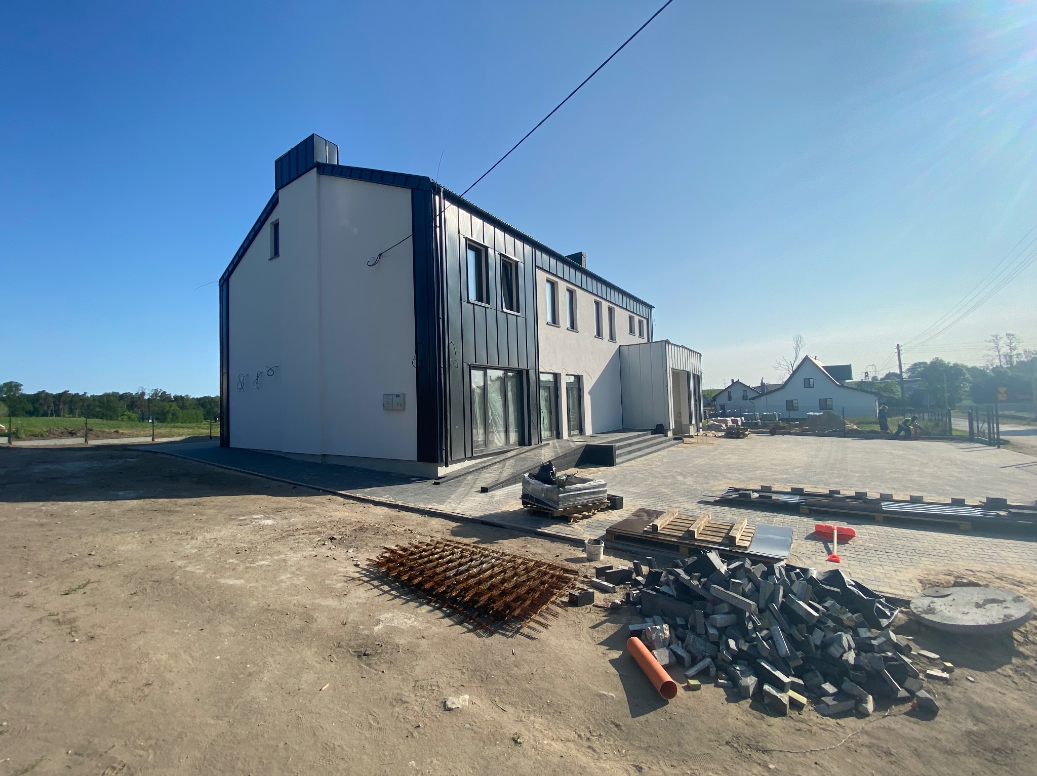 Nowoczesny, dwukondygnacyjny budynek z grafitowym dachem i elementami elewacji. Na pierwszym planie materiały budowlane: kostka brukowa i zbrojenie.