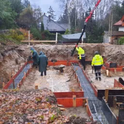 Wykop pod fundamenty domu jednorodzinnego z widocznym szalunkiem, zbrojeniem i ekipą budowlaną w trakcie wylewania betonu za pomocą pompy, w tle dom w stanie surowym.
