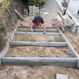 Pracownik w niebieskich rękawicach układa stopnie schodów z szarej kostki betonowej, widoczne narzędzia i materiały budowlane na placu budowy.