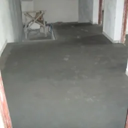 Świeżo wylana, szara podłoga w pomieszczeniu w trakcie remontu, widoczne ślady butów na powierzchni, drewniany kozioł i wiadro w tle.