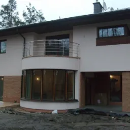 Nowoczesny dom jednorodzinny z elewacją w jasnym kolorze, fragmentami wykończonymi cegłą, z balkonem z metalową balustradą, na tle lasu.