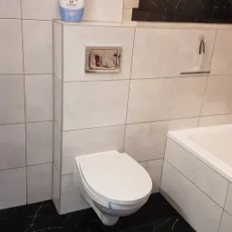 Wykończona łazienka z białymi płytkami imitującymi beton, czarną marmurową podłogą, podwieszaną toaletą i ukrytą spłuczką. Widoczna również wanna oraz pralka.