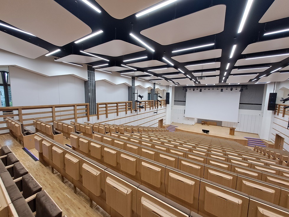 Wnętrze nowoczesnej auli z drewnianymi ławkami i okładzinami ściennymi HPL, podwieszany sufit z oświetleniem liniowym i sceną w tle.