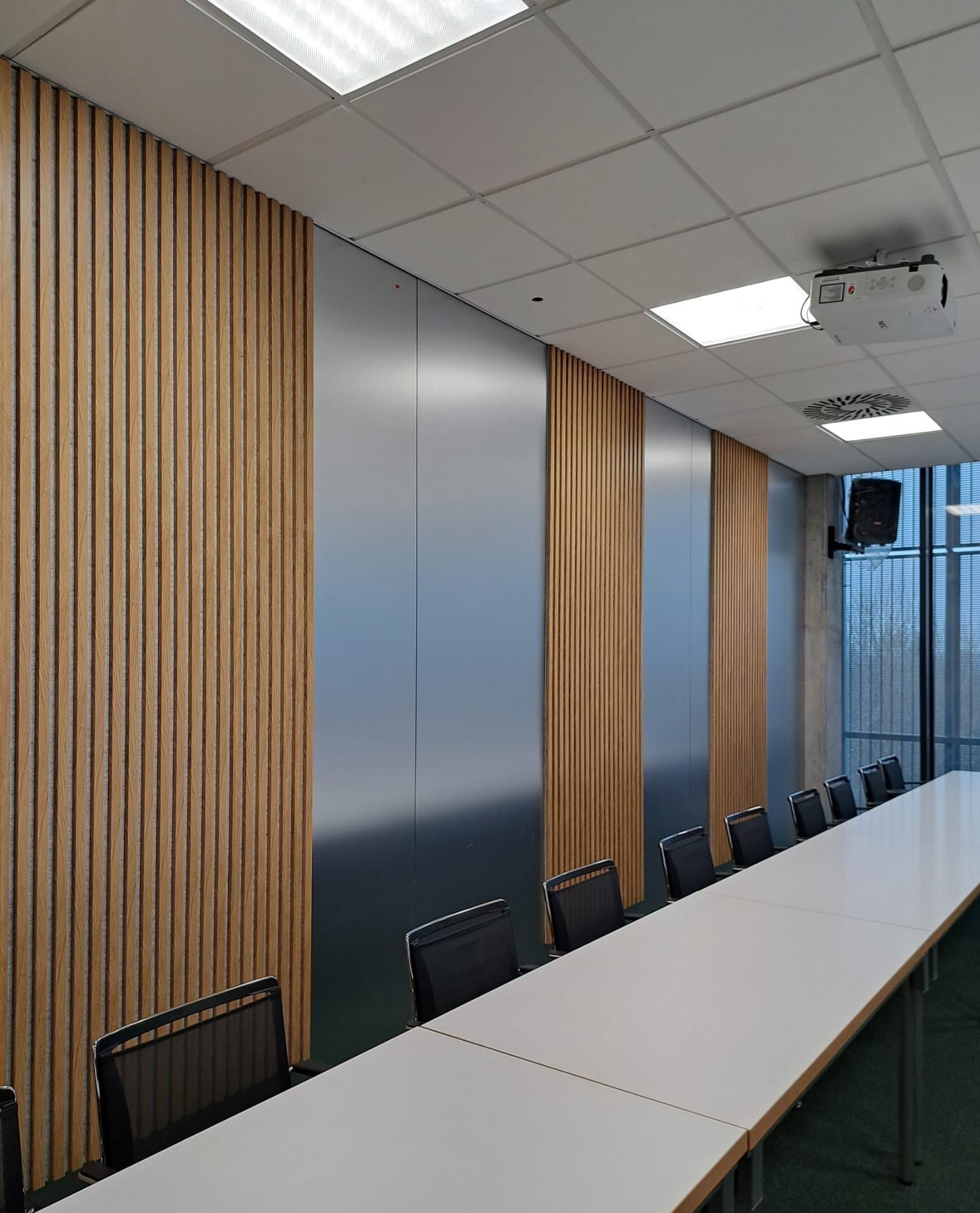 Sala konferencyjna z okładzinami ściennymi HPL w odcieniach drewna i szarości. Widoczne krzesła, stół, projektor i kratka wentylacyjna na suficie.