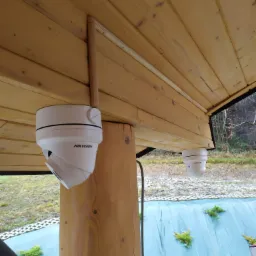 Dwie kamery kopułkowe Hikvision zamontowane pod drewnianym zadaszeniem, widok z dołu, na zewnątrz widoczna zielona skarpa z roślinnością i drabina.