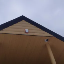 Kamera monitoringu zamontowana pod drewnianym okapem dachu, widoczny także sygnalizator alarmowy i drewniane słupy podtrzymujące dach.