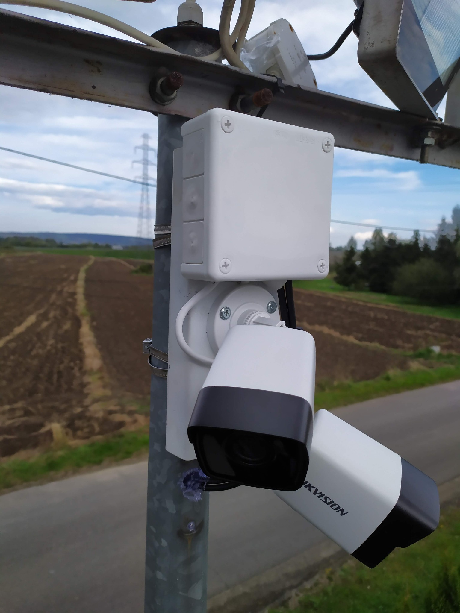 Zewnętrzna kamera monitoringu Hikvision przymocowana do metalowego słupa na tle pola uprawnego i linii wysokiego napięcia.