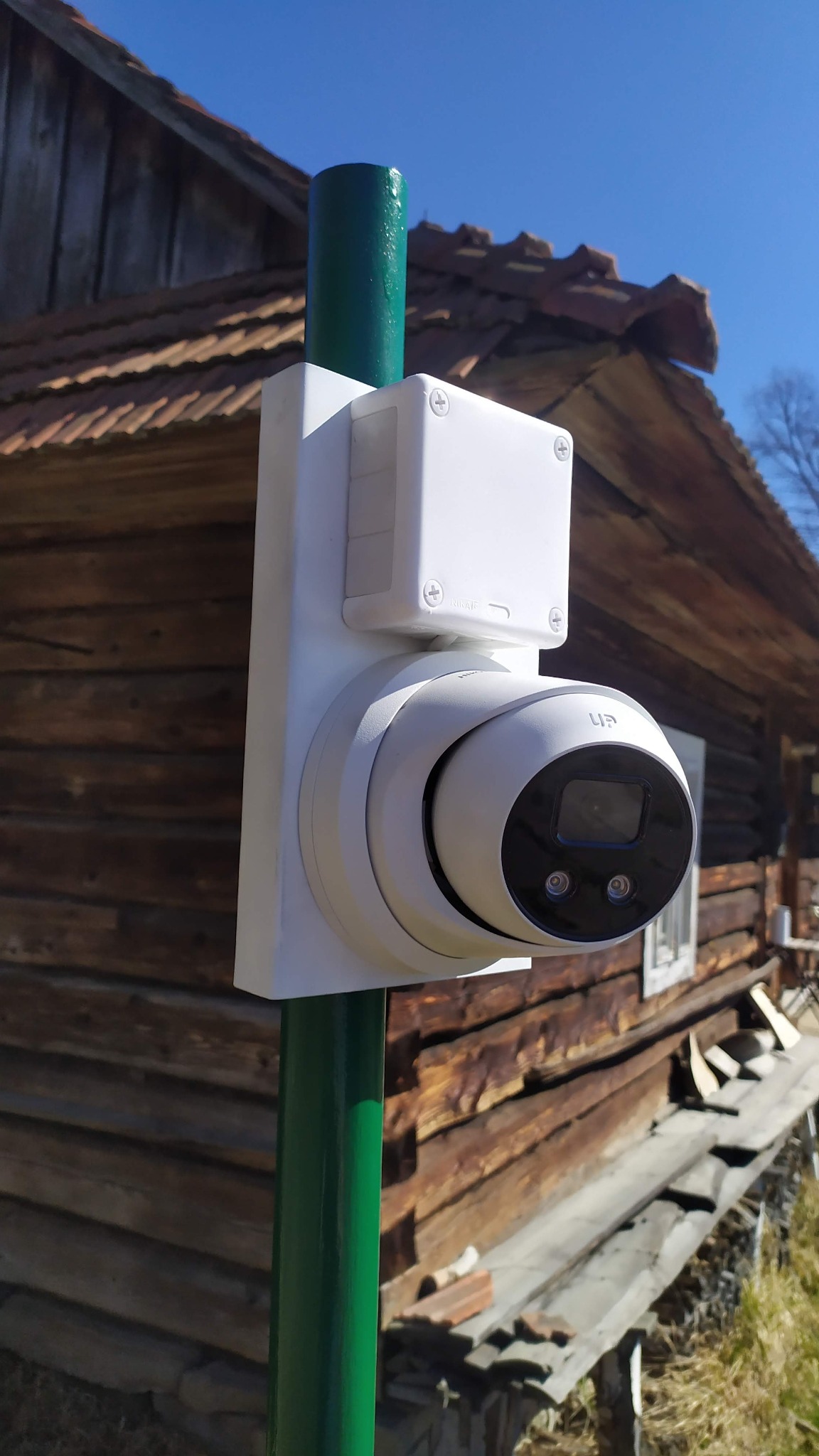 Kamera monitoringu zamontowana na zielonym słupku przed drewnianym domem w Nowym Sączu, widoczne mocowanie kamery i puszka instalacyjna.