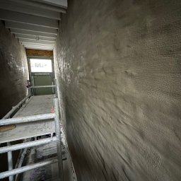 KS Kajetan Sobel - Wąskie wnętrze w trakcie prac wykończeniowych z nałożoną szarą warstwą na ścianach, widoczne rusztowanie i okno na końcu korytarza.