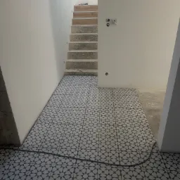 Ułożona, wzorzysta posadzka w przedpokoju z betonowymi schodami i gładkimi, białymi ścianami. Widoczne niedokończone prace na ścianach i podłodze.