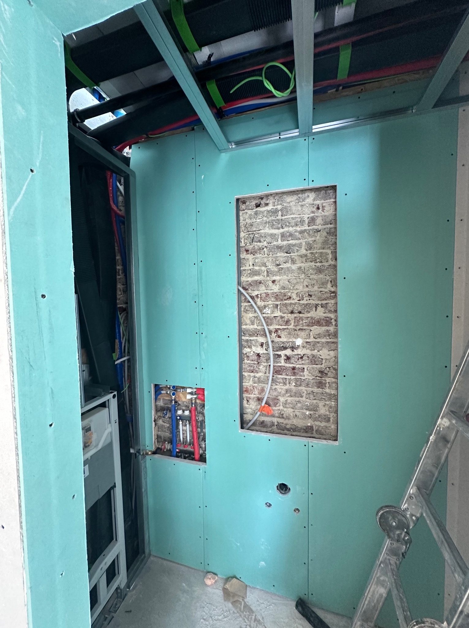 Montaż zielonych płyt gipsowo-kartonowych na ścianie z widoczną instalacją hydrauliczną i fragmentem muru z cegły w otworze